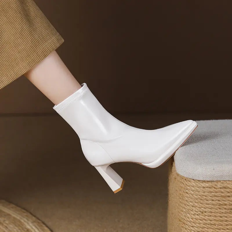 White patent leather ankle boots with a pointed toe and sculptural block heel, shown on a leg with a tan corduroy pant.