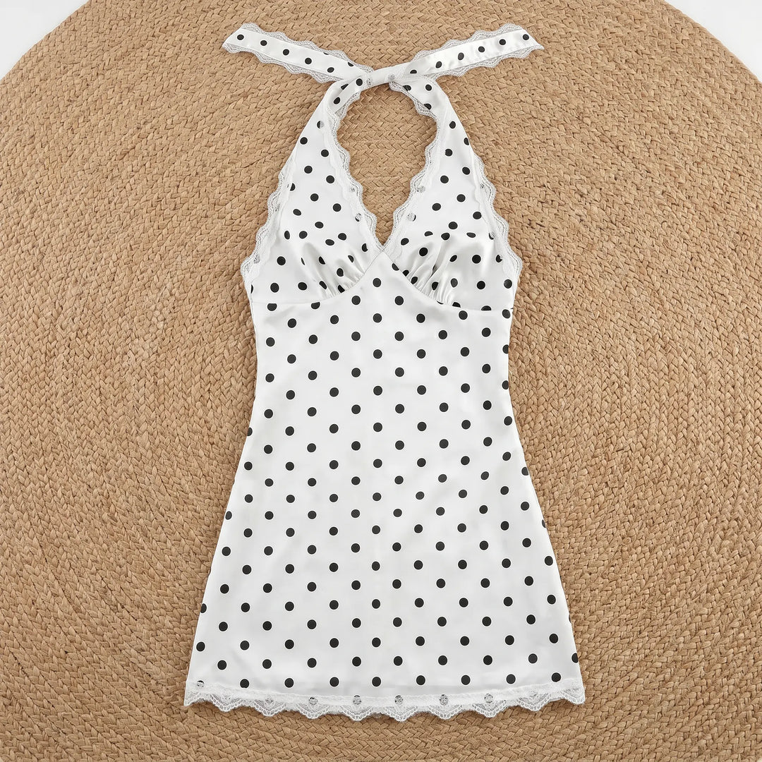 White mini halter neck dress with black polka dots, featuring delicate white lace trim on the V-neckline and hem, laid flat on a woven background.