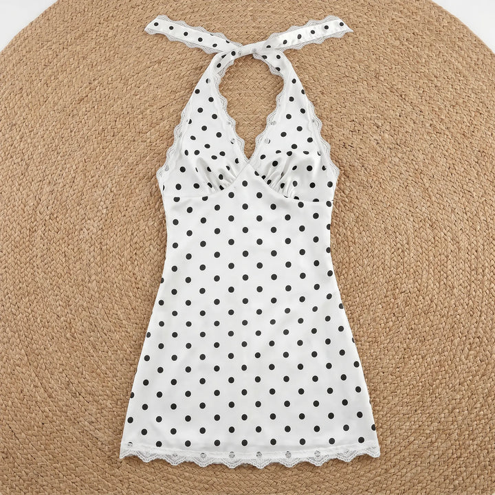 White mini halter neck dress with black polka dots, featuring delicate white lace trim on the V-neckline and hem, laid flat on a woven background.