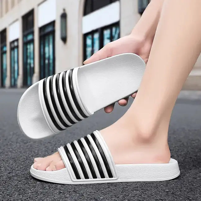 Person wearing white EVA comfort slides with black signature stripes, shown from the side outdoors on a street.