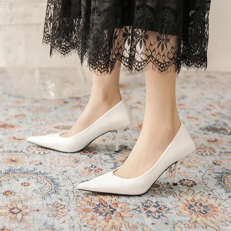 White high-gloss faux leather stiletto heels featuring a sharp pointed toe, low-cut vamp, and a unique sculpted heel base, shown on a model's foot paired with a black lace skirt.