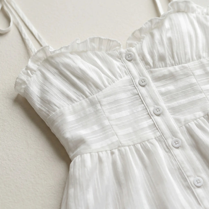 Close-up of a white cotton voile mini dress featuring a sweetheart neckline with delicate ruffles, adjustable spaghetti straps, and functional pearlized button-front detailing.