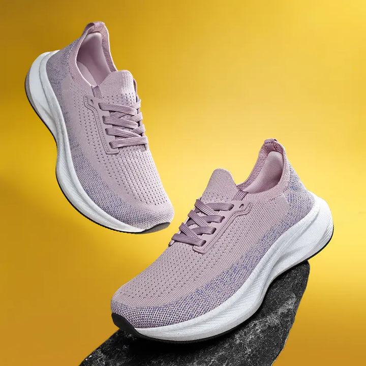 Pair of dusty rose wide fit breathable athletic trainers with white soles, shown from a three-quarter angle on a textured surface against a yellow gradient background.