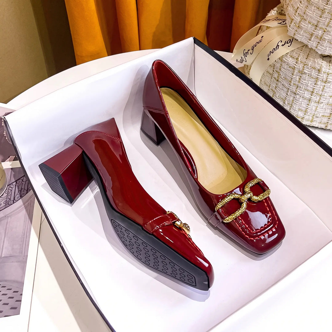 Pair of wine red patent leather square toe pumps with gold buckle embellishment and block heels, presented in a studio setting.