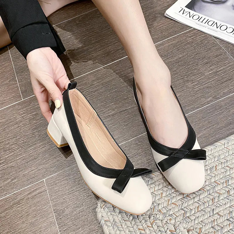 Women's beige square-toe ballet flats with a black satin bow accent and contrasting black trim, shown worn on feet with a low block heel.