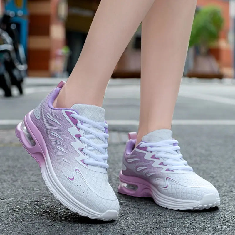 Women's breathable running trainers in gradient purple and pink, worn on feet with a visible air cushion heel, showcasing their lightweight, sporty style on an outdoor street.