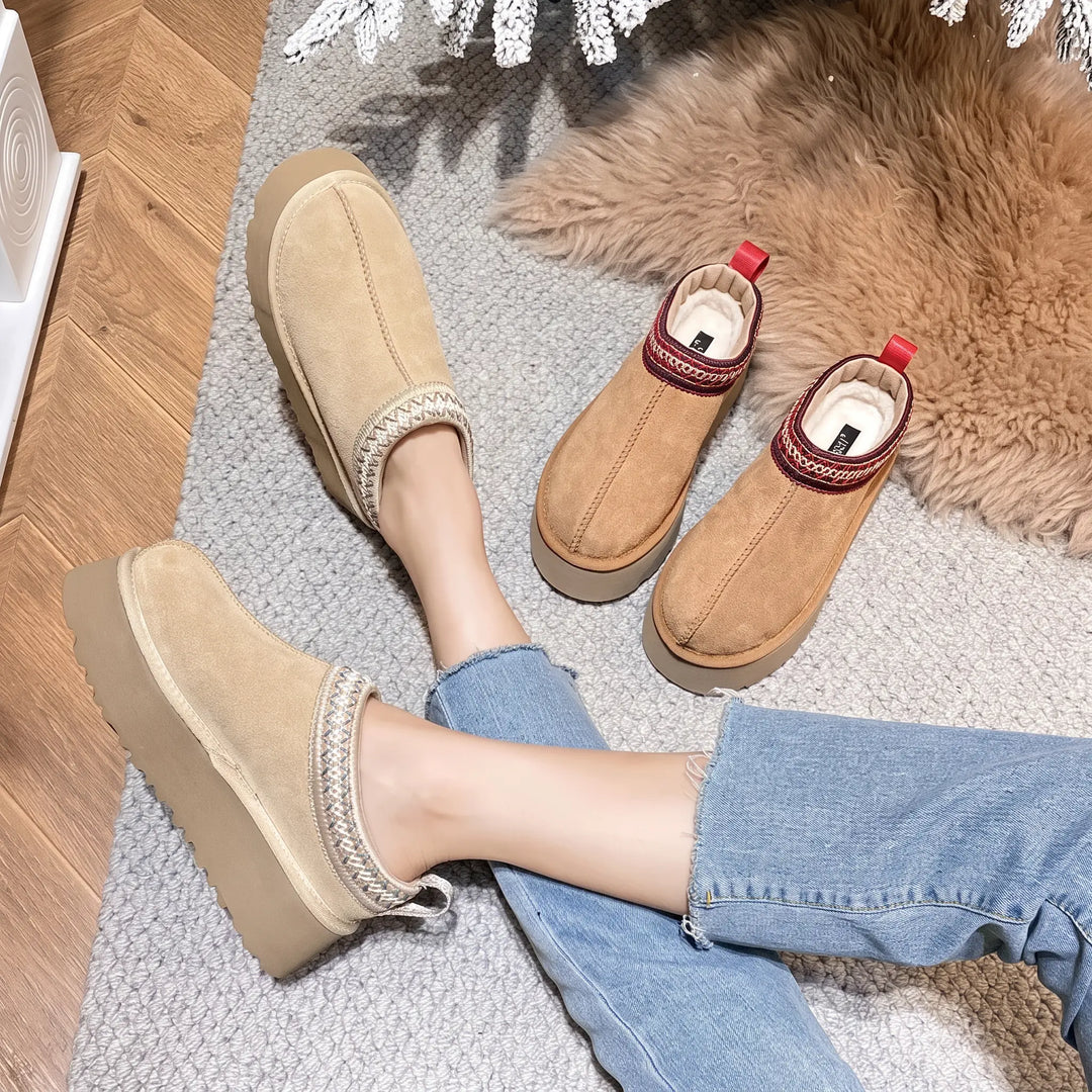 Women wearing cozy tan faux suede slip-on boots with plush white faux fur lining and a decorative red, white, and beige woven band, resting on a rug. These casual winter footwear feature a thick platform sole and a red heel pull-tab.