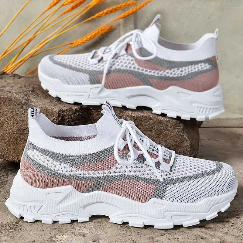Women's white mesh sneakers with pink and grey abstract patterns, featuring a sock-like collar and a sculpted chunky white sole, displayed on a textured stone surface with dried wheat stalks.