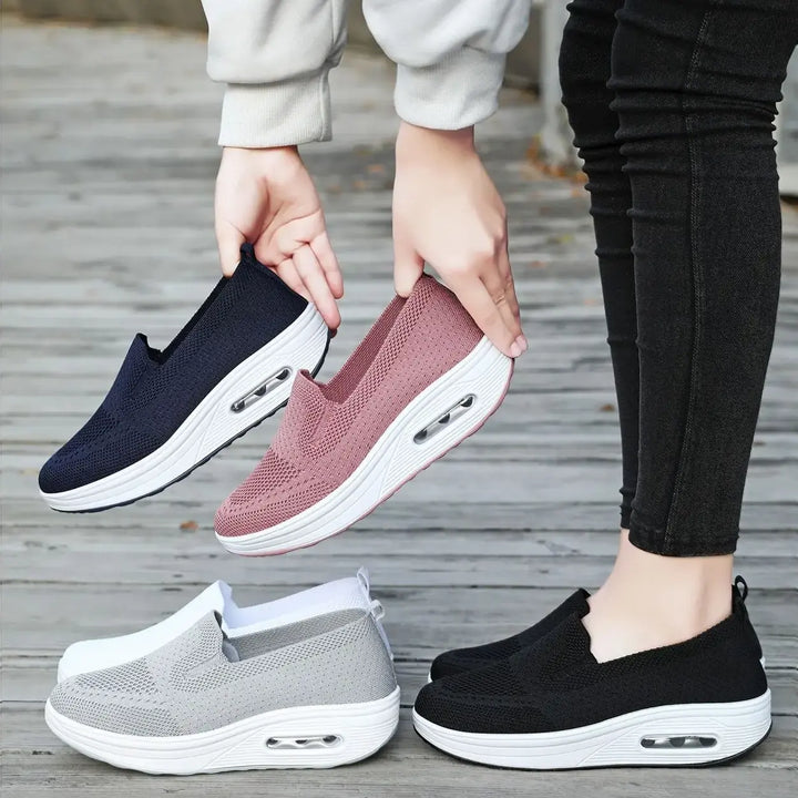 Showcasing multiple colors of Xylos breathable knit slip-on sneakers with air cushion soles, including navy, pink, grey, white, and black, with a foot modeling the black pair on a wooden surface.