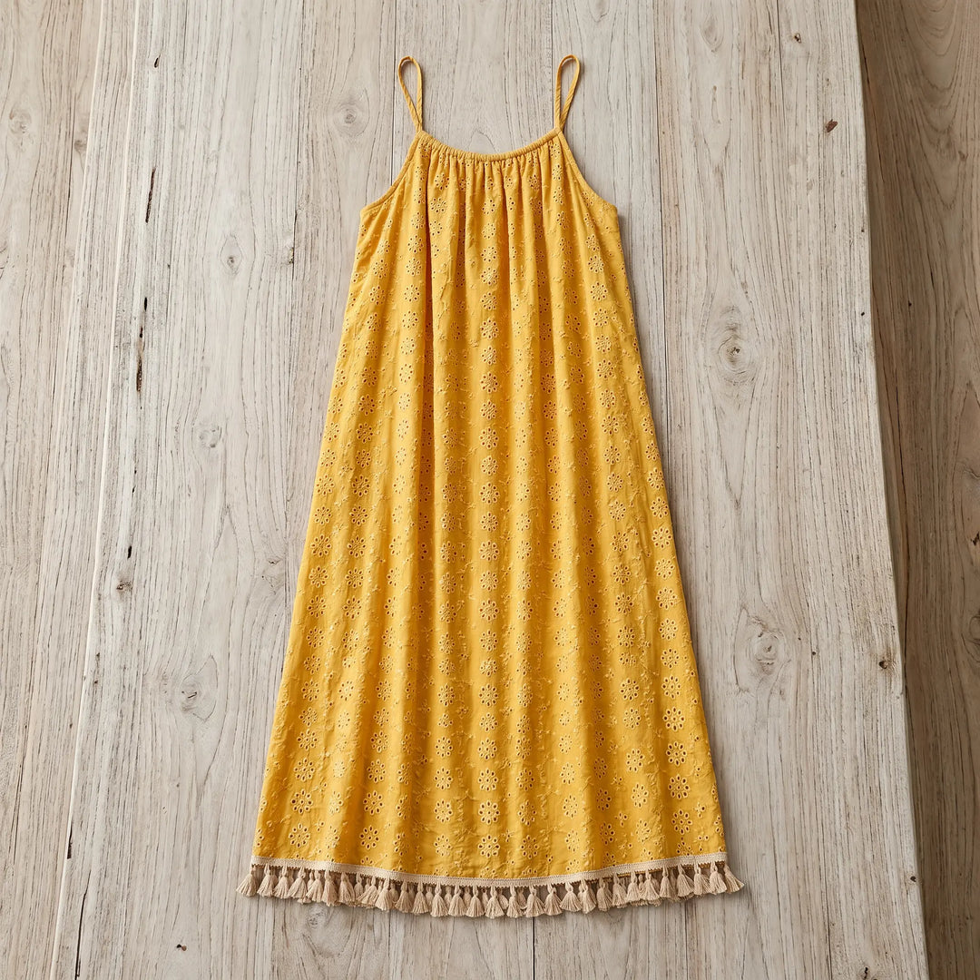 Flat lay of a golden yellow bohemian maxi dress featuring all-over tonal embroidery and a white tassel hem, displayed on a rustic wooden surface.