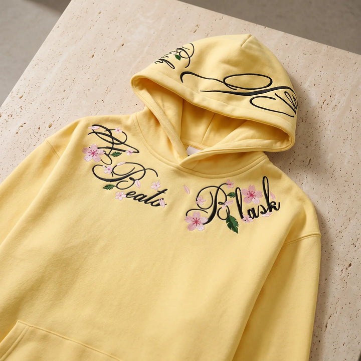 Soft yellow Orvela relaxed-fit hoodie featuring intricate floral and script embroidery around the neckline and on the hood, shown at a slight front angle.