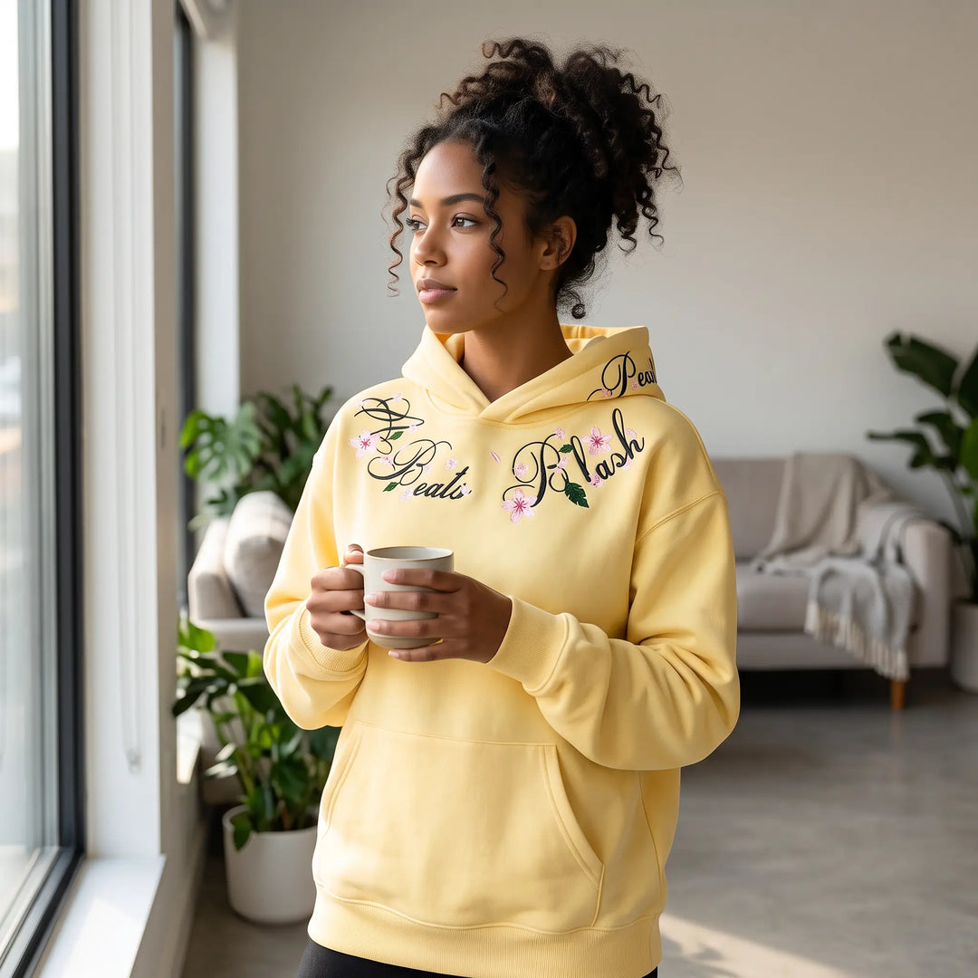 A young woman stands by a window holding a mug, wearing a relaxed-fit, pale yellow hoodie with intricate floral embroidery around the neckline. The lifestyle shot captures a cozy, casual morning vibe.