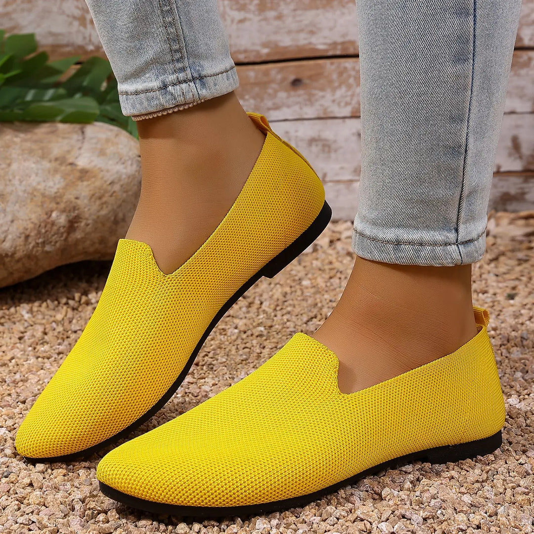 Vibrant yellow knitted pointed toe flats worn by a person with light blue jeans, showcased in a lifestyle setting with pebble ground and rustic wooden wall backdrop.