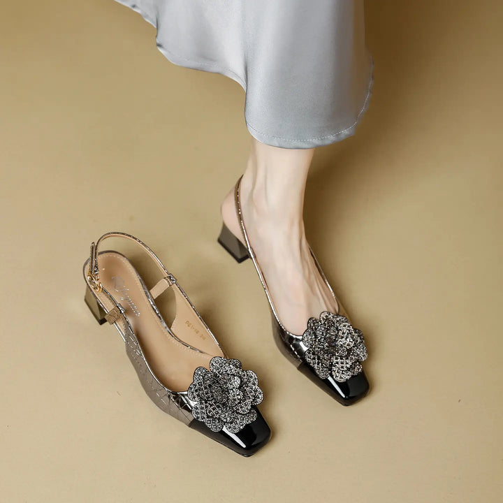 Silver metallic slingback heels featuring a sparkling jeweled floral appliqué on the vamp, a glossy black patent cap toe, and a chunky block heel, styled with a flowing satin skirt.