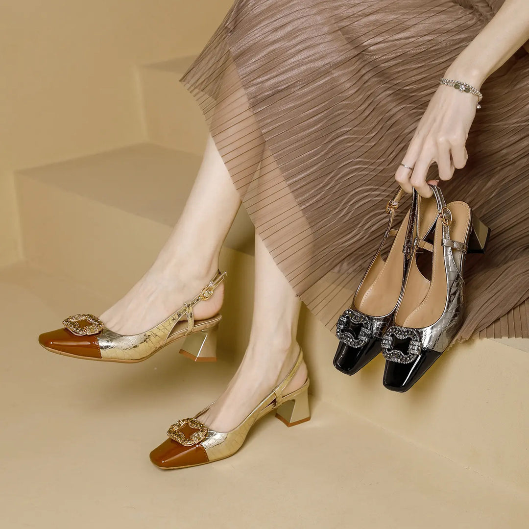 Close-up of a woman wearing gold metallic slingback heels with a patent brown toe cap and crystal-encrusted square buckle, while holding a gunmetal pair.