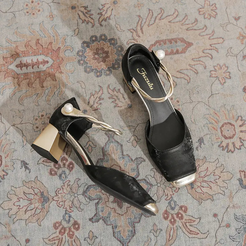 Pair of black Zephyra chunky heel sandals with gold-capped square toes and a braided ankle strap featuring a faux pearl accent, displayed on a patterned rug.