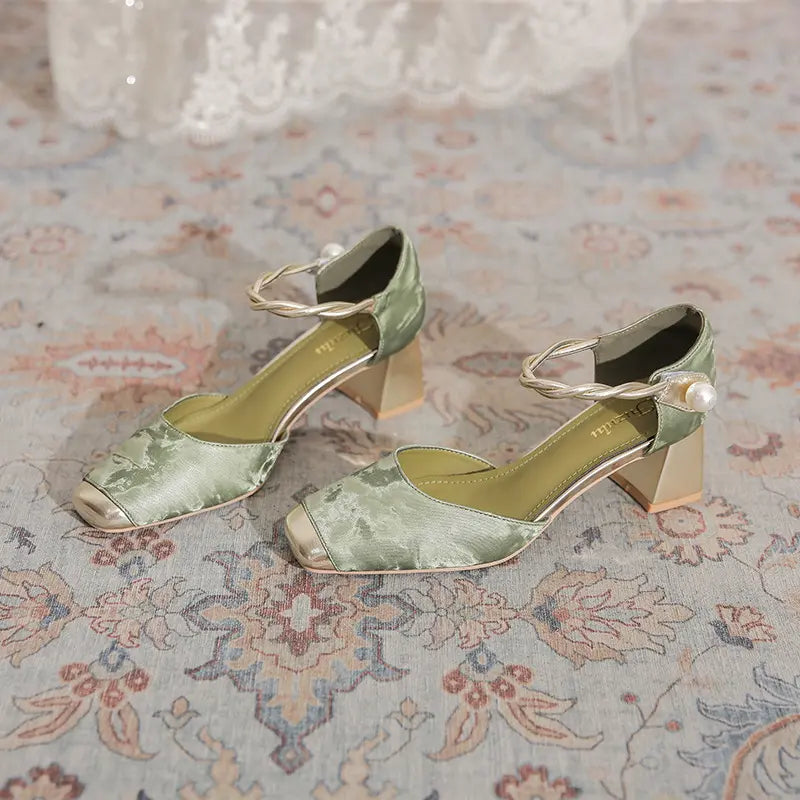 Elegant sage green Zephyra chunky heel sandals featuring a square toe with a gold metallic cap, braided ankle strap with faux pearl, and a geometric block heel, displayed on a patterned rug.