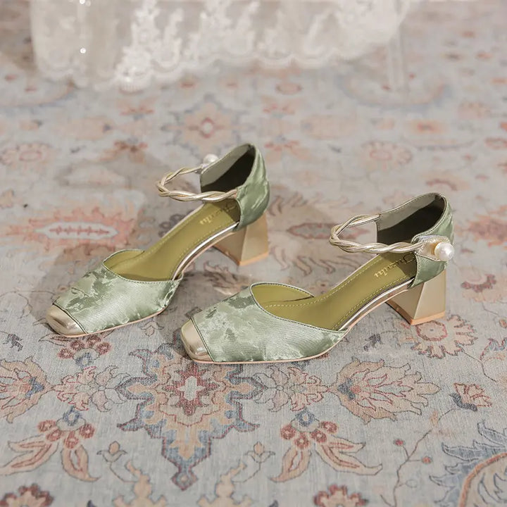 Elegant sage green Zephyra chunky heel sandals featuring a square toe with a gold metallic cap, braided ankle strap with faux pearl, and a geometric block heel, displayed on a patterned rug.