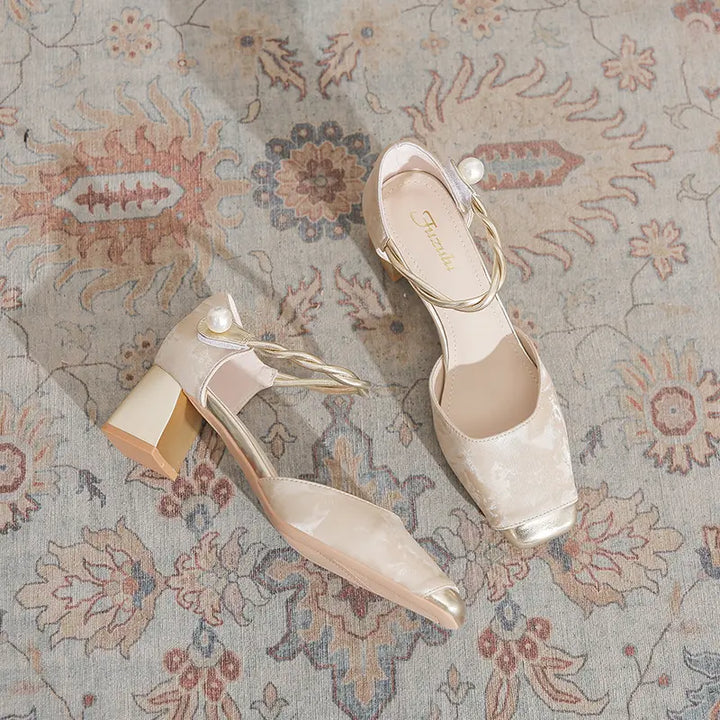 A pair of elegant champagne beige Zephyra chunky heel sandals with gold-capped square toes and braided pearl ankle straps, artfully displayed on a textured rug.