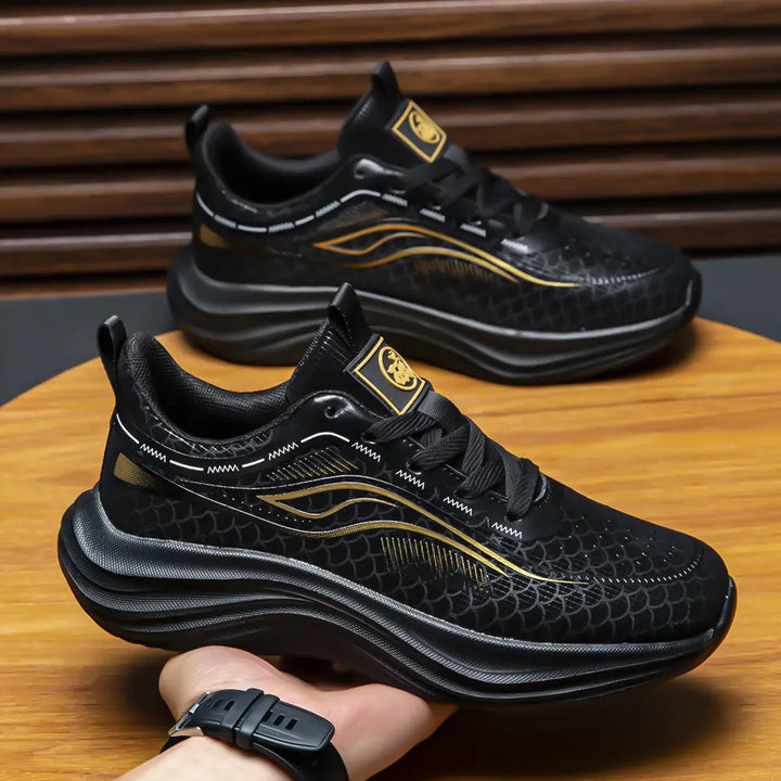 Black Zephyrin performance sneakers with metallic gold wave graphics and scaled pattern on the mesh upper, shown from a three-quarter angle with a sculpted black sole. The shoe features a white lace-up closure and a padded heel collar. A hand wearing a black watch holds the shoe against a studio background.