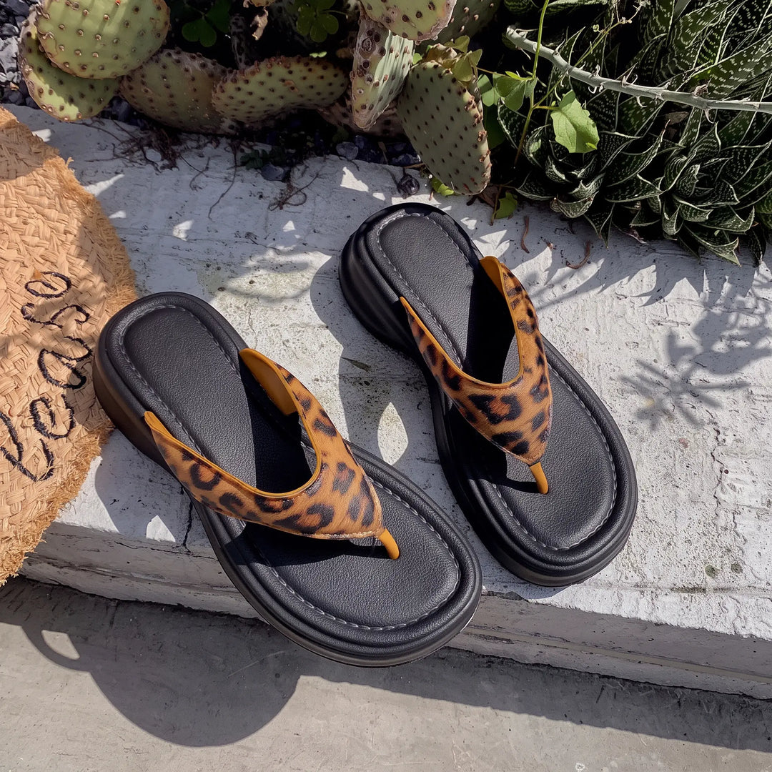 A pair of Zethel platform thong sandals featuring a leopard print upper strap and a thick, matte black EVA sole, styled casually with a straw bag and greenery.