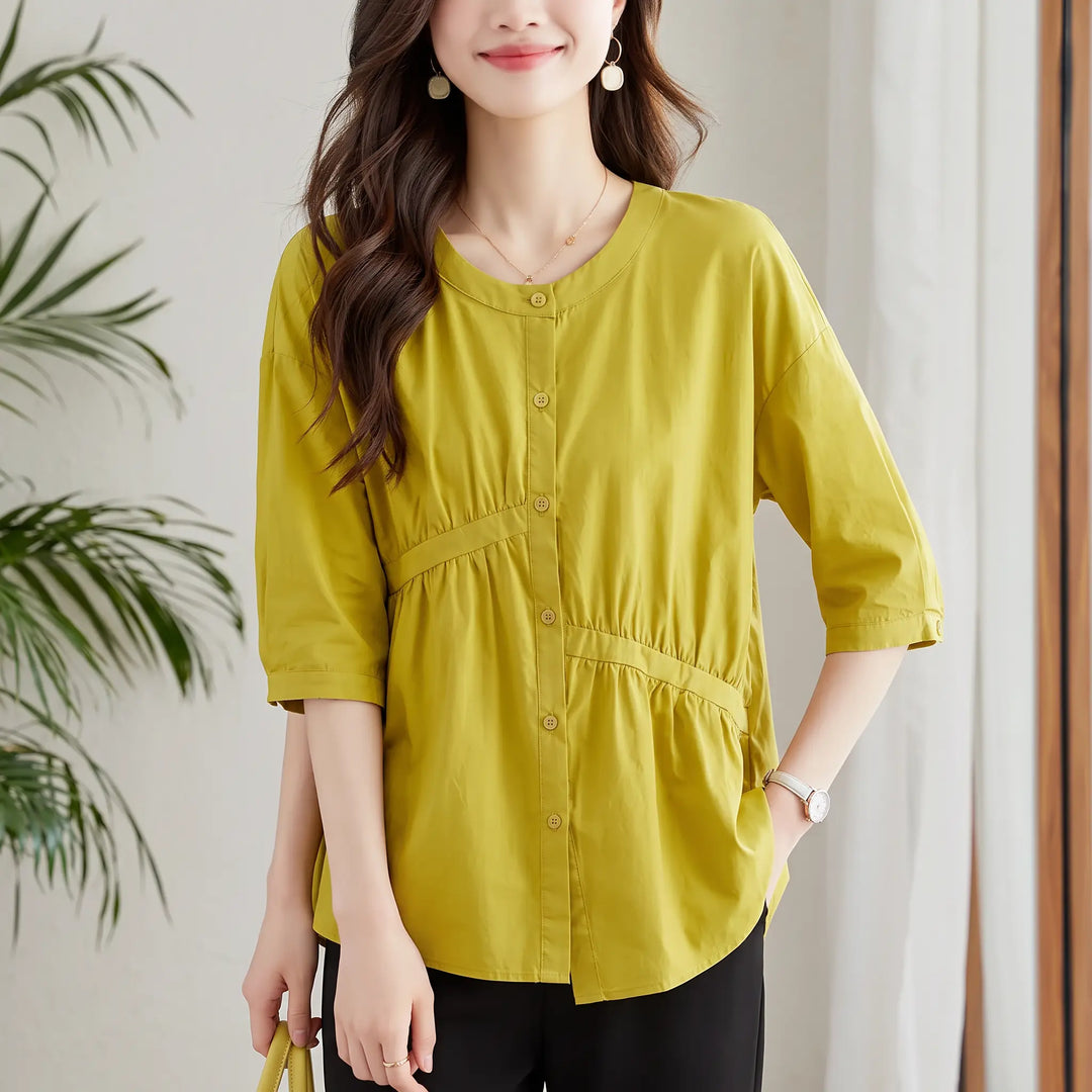 A woman wearing a Zolani chic pleated cotton top in warm mustard green, featuring a round neckline, diagonal waist pleating, and asymmetric peplum hem.