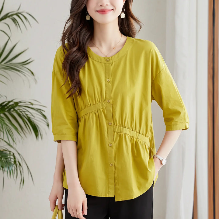 A woman wearing a Zolani chic pleated cotton top in warm mustard green, featuring a round neckline, diagonal waist pleating, and asymmetric peplum hem.