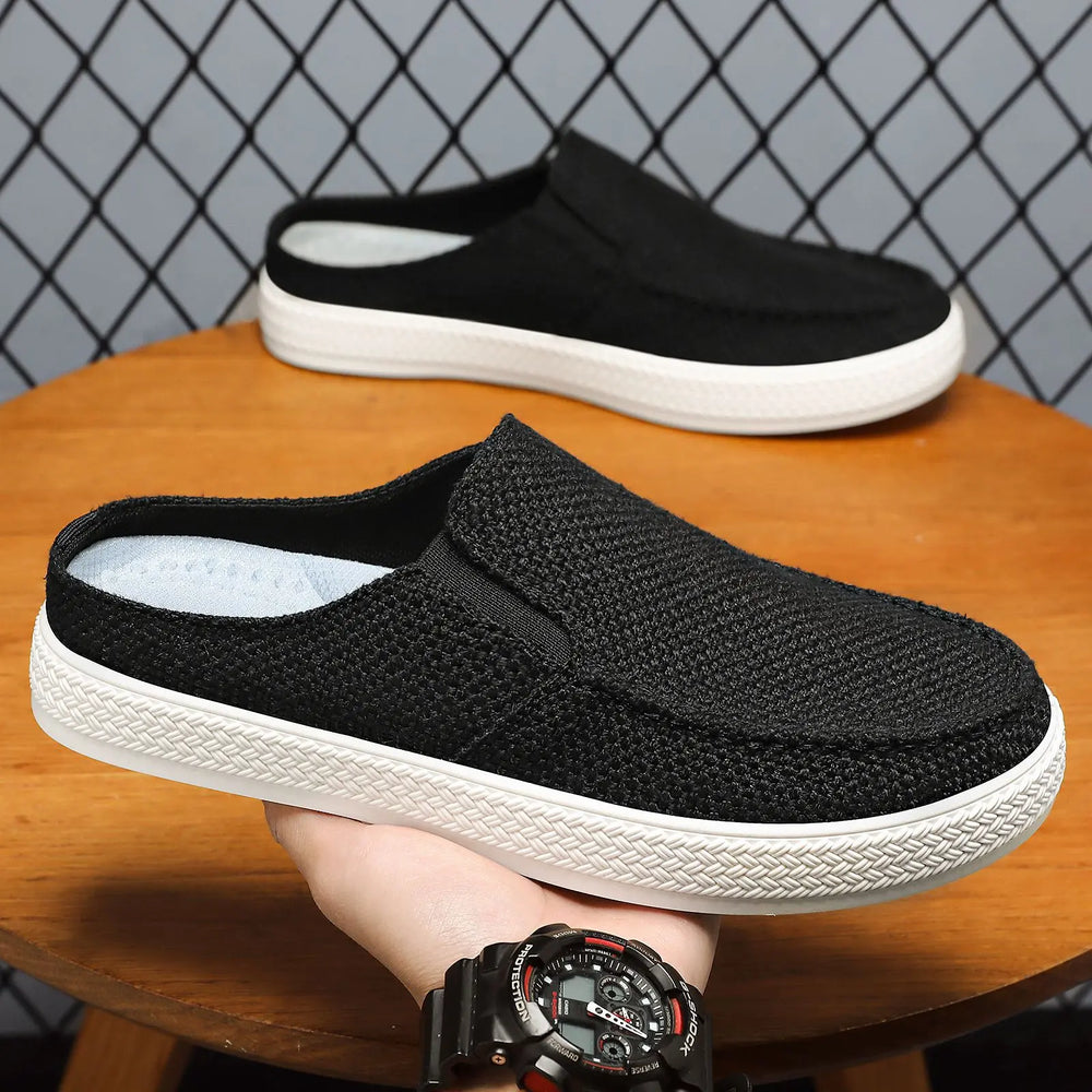 Men's black breathable canvas backless mules shown in a three-quarter view, held by a hand with a watch against a textured grey background.