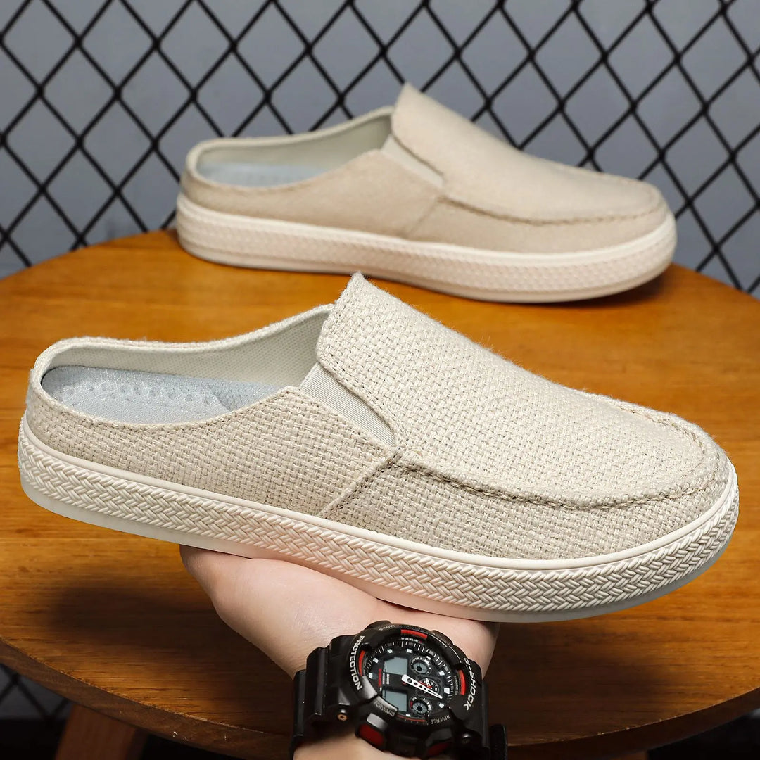 Men's Zoltan breathable canvas mules in oat beige, shown from a three-quarter angle on a wooden surface with a second pair in the background.
