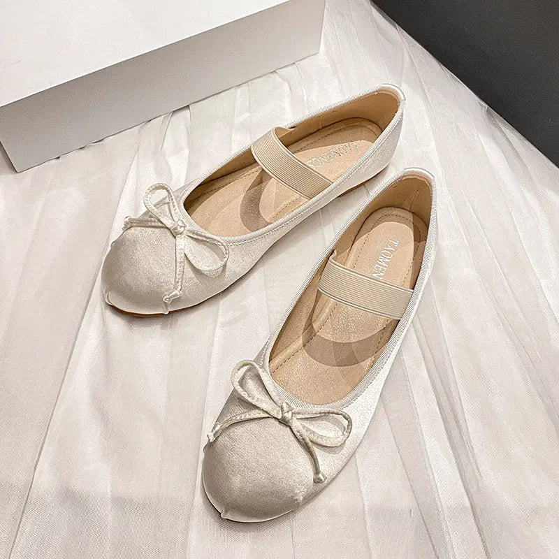 A pair of Ivory White satin ballet flats with delicate bow details and elastic instep straps, displayed flat lay on a draped white fabric background.