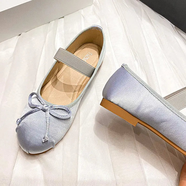 Pair of Zoryan satin ballet flats in light blue, featuring a delicate bow on the vamp and an elastic instep strap, shown from a three-quarter angle on a soft white background.