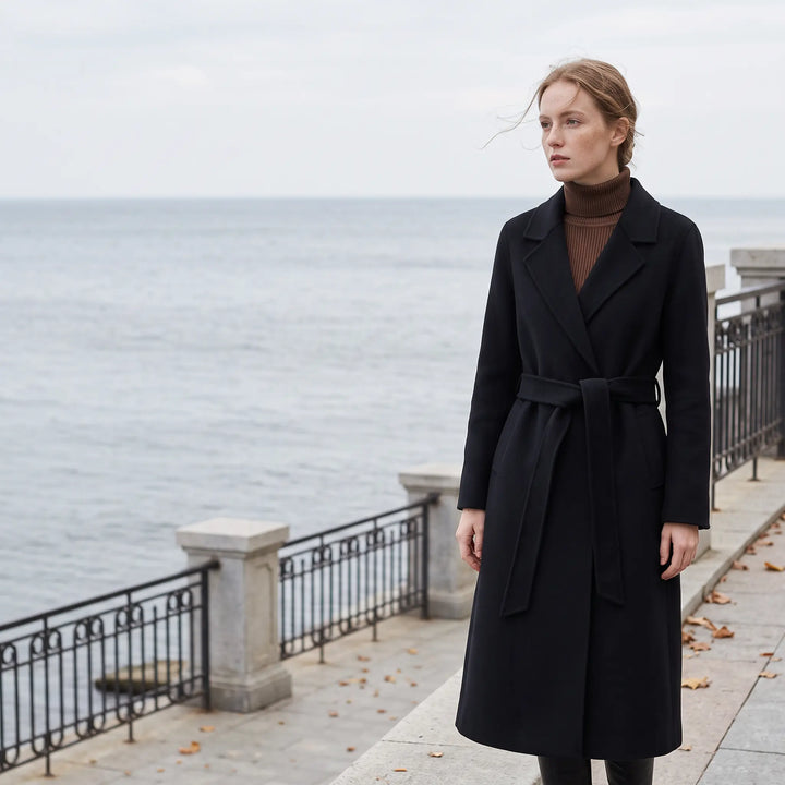 A woman wearing a black Zoryn tailored wool coat with a self-tie belt, standing outdoors by the water on a grey day.