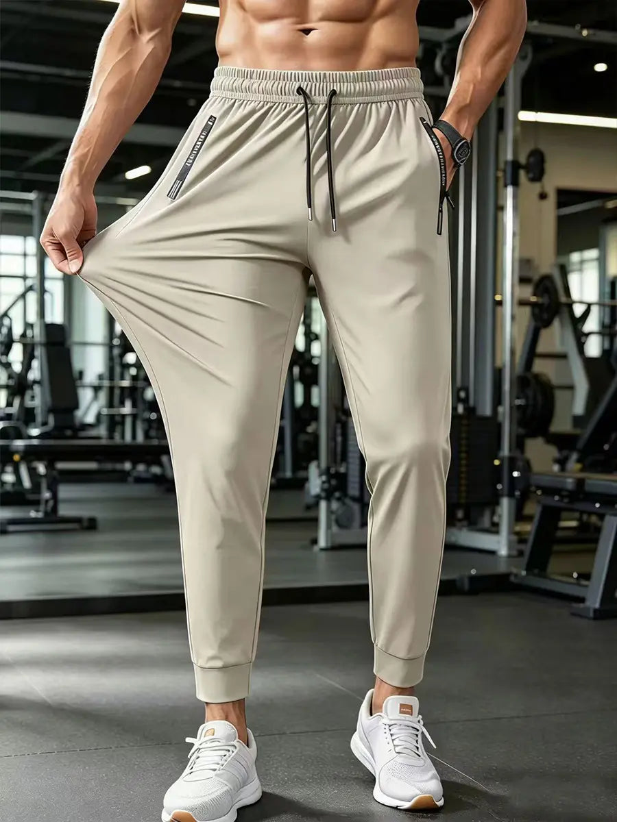 Men's lightweight Zydron trousers in light khaki, shown on a model demonstrating high stretch fabric in a gym setting with zippered pockets and cuffed ankles.