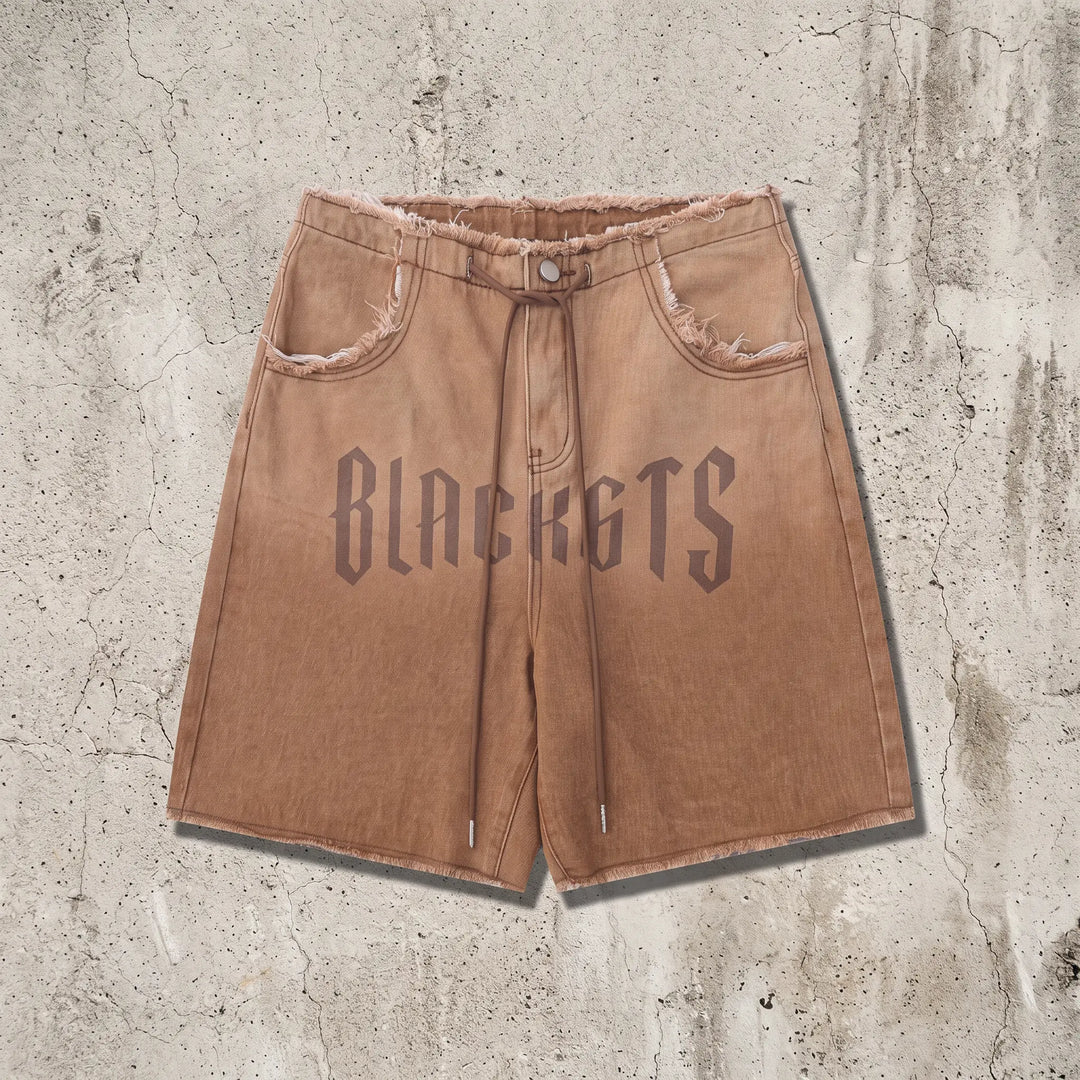 Zykra wide leg denim shorts in gradient wash from brown to dusty rose, featuring distressed frayed hem, drawstring waist, and bold 'BLACKGTS' graphic print on the leg.