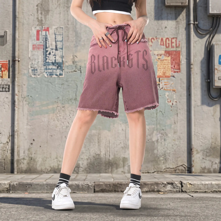 Zykra wide-leg denim shorts in ombré brown to dusty rose wash, featuring a frayed hem, drawstring waist, and large BLACKGTS graphic print on the leg. Front view studio shot.