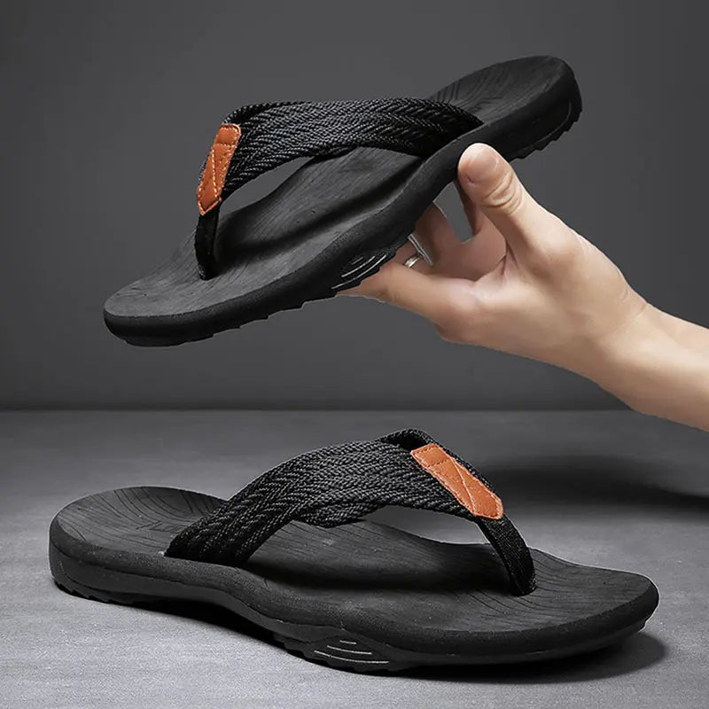 Men's Zylar casual flip-flops in black, featuring braided textile straps and a contoured EVA footbed, displayed with a hand holding one sandal above another on a studio background.