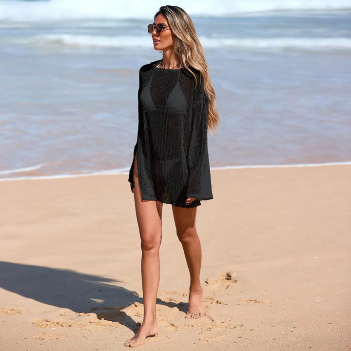 Woman walking on a sandy beach wearing a black sheer metallic knit Zylia beach cover-up with bell sleeves and side slits over a bikini.