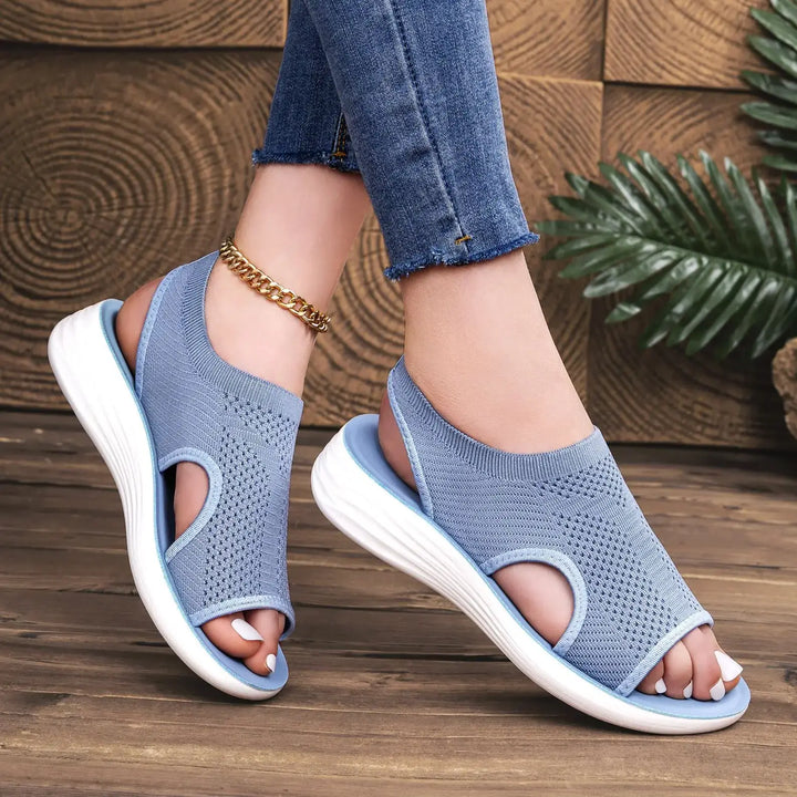 Muted blue Zyloria knit sandals with an open-toe design and a geometric weave, worn by a model in jeans with a gold anklet, set against a rustic wood and greenery background.