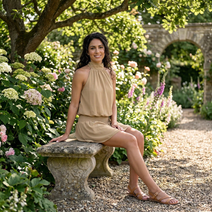A model wears a flowy beige halter neck mini dress with a shirred waist, sitting on a stone bench in a lush garden setting.