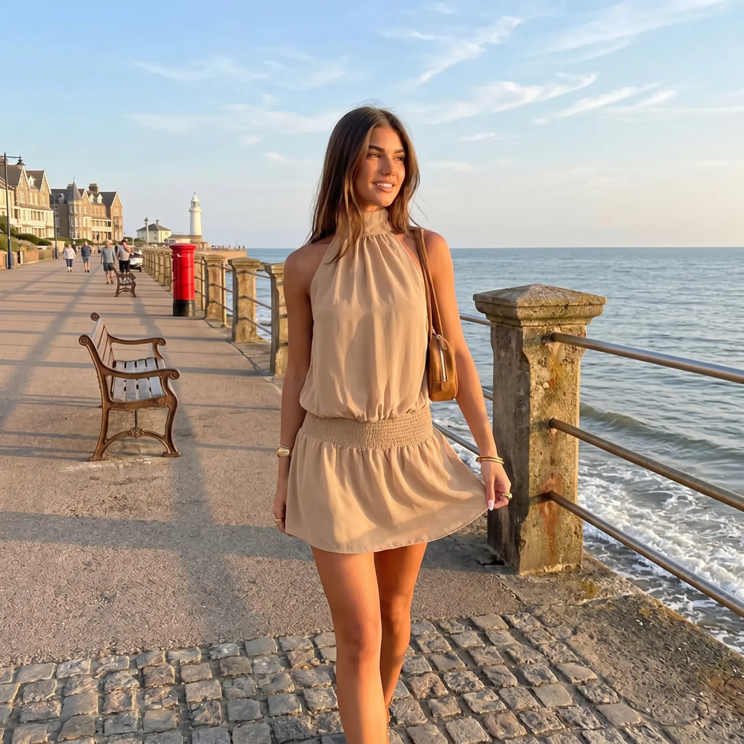 A young woman walks along a seaside promenade wearing a flowing beige halter neck mini dress with a shirred waist. She carries a small brown bag and wears gold bracelets.