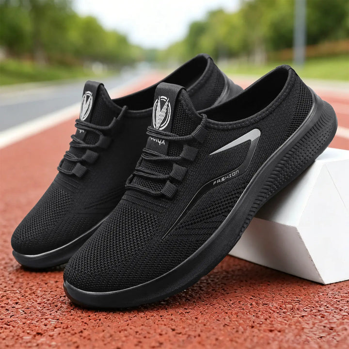 Three-quarter view of black Zylos breathable comfort slip-on trainers with elastic laces and a reflective side stripe that reads &