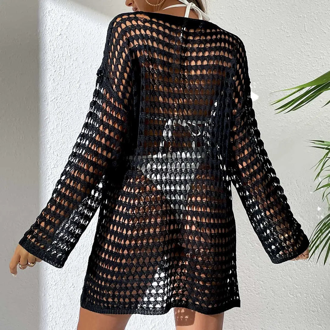Back view of a black crochet knit beach cover-up dress with long sleeves, worn over white swimwear in a resort setting with a palm leaf.