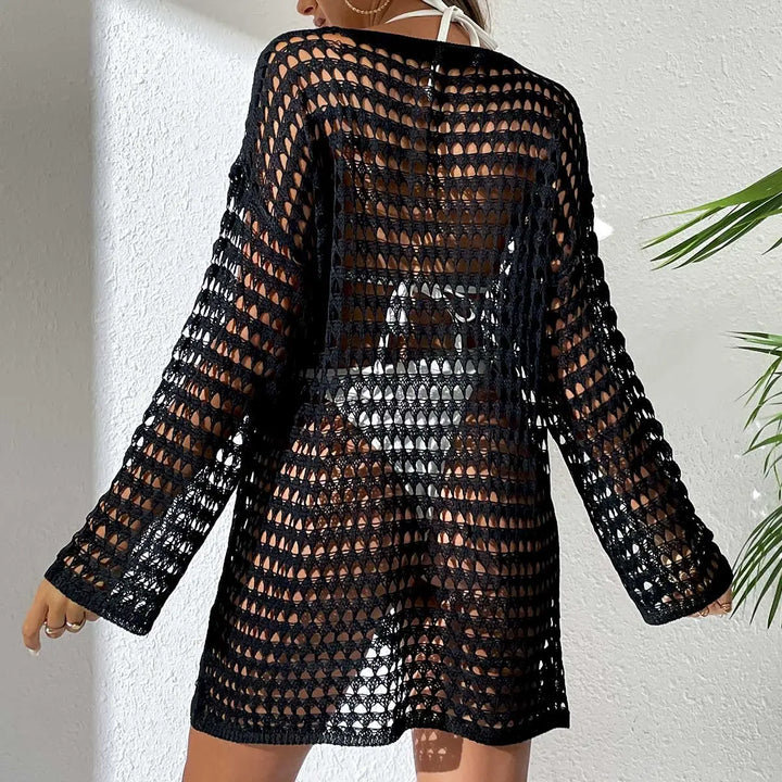 Back view of a black crochet knit beach cover-up dress with long sleeves, worn over white swimwear in a resort setting with a palm leaf.