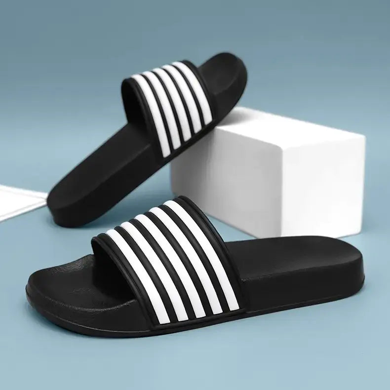 A pair of black Zylos EVA Comfort slides with white stripe detailing on the strap, shown on a studio background with a white block prop.
