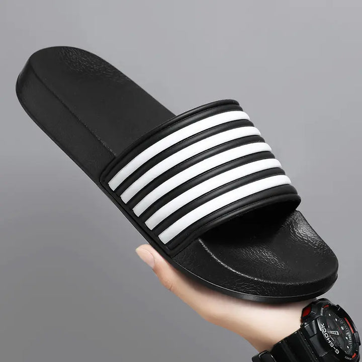 A Zylos black EVA comfort slide sandal featuring a wide strap with iconic white diagonal stripes, held by a hand against a studio background.