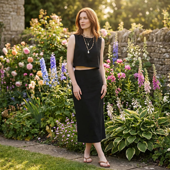 A woman models a black linen-blend sleeveless crop top and fitted midi skirt set in a vibrant English garden setting, showcasing its elegant, flowing silhouette.
