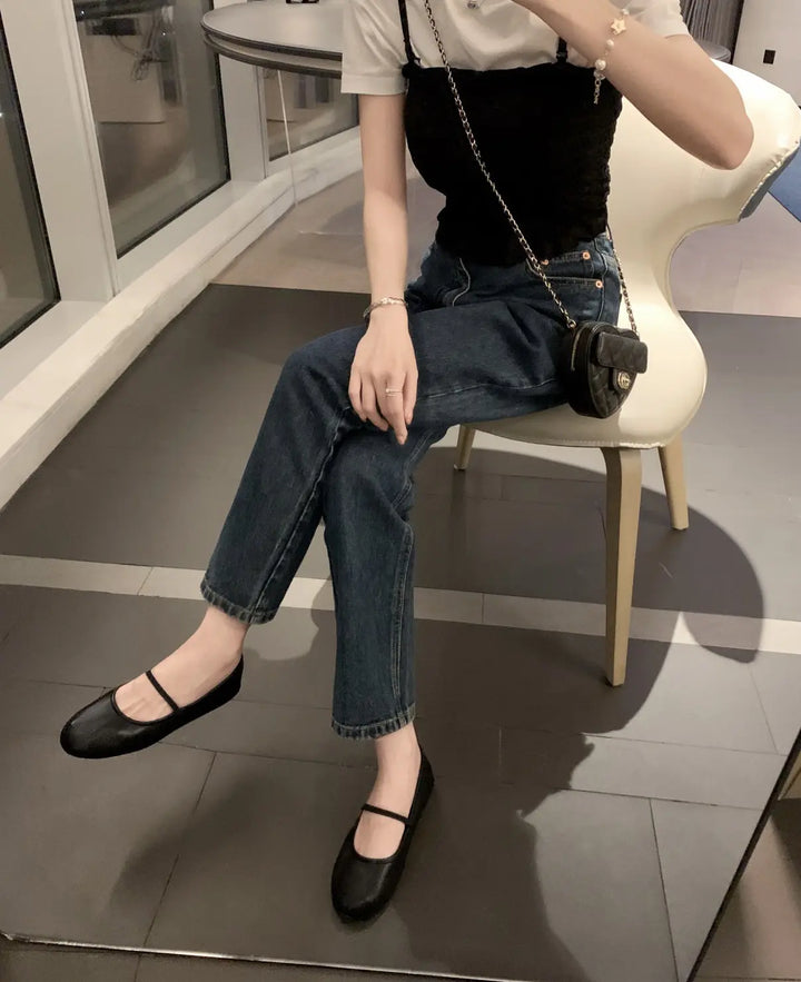 A woman wearing black Zylos ballet flats with a minimalist Mary Jane strap, paired with dark wash jeans and a black top while sitting on a chair.