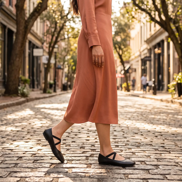 Chic black Mary Jane ballet flats with elastic straps worn by a woman walking on a cobblestone street, paired with a flowing terracotta dress.