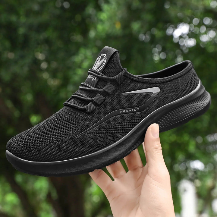 A black Zylos breathable mesh slip-on trainer held by a hand, showcasing its side profile, elastic laces, and textured sole against a blurred natural green background.