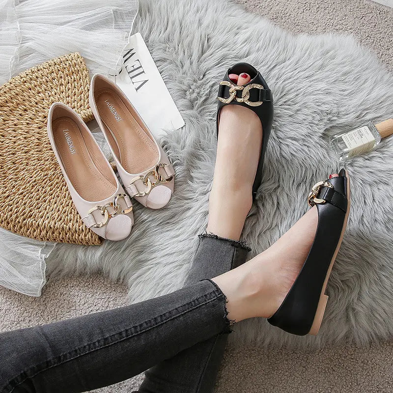 A pair of black peep-toe flat shoes with gold chain detailing on the vamp, worn by a person in dark jeans on a grey rug, alongside a pair of cream flats.